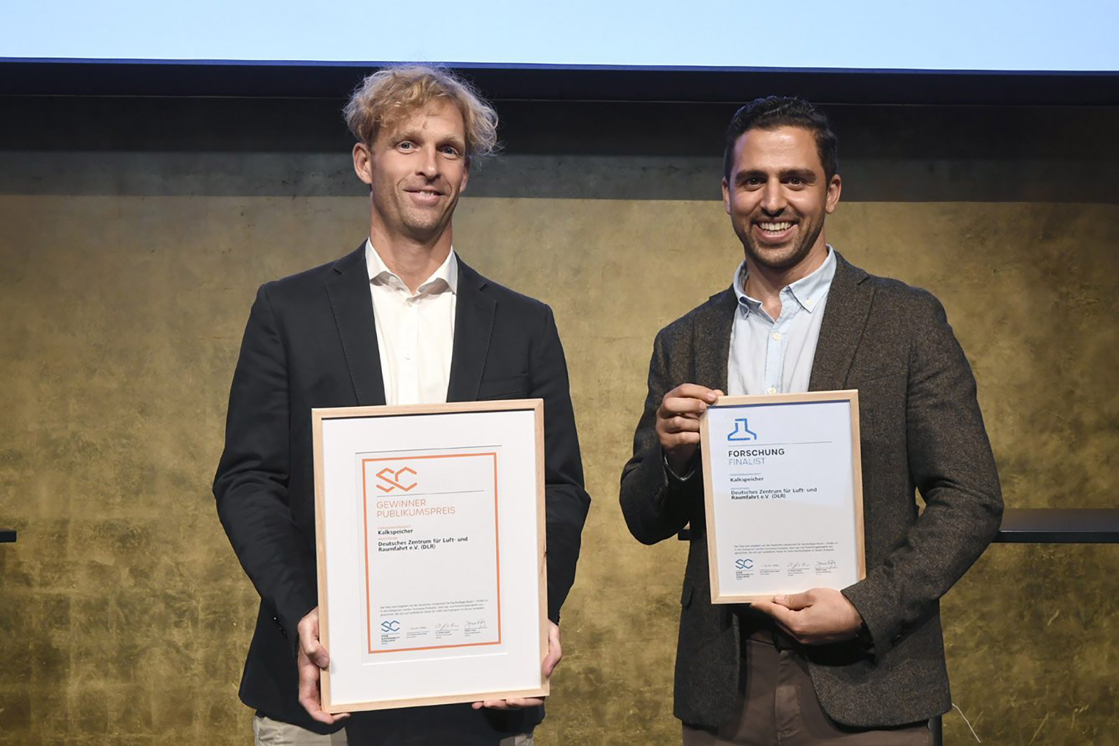 Zwei junge Herren sind die Gewinner der DGNB Sustainability Challenge 2023. Sie halten Urkunden in der Hand und zeigen diese lachend in die Kamera.