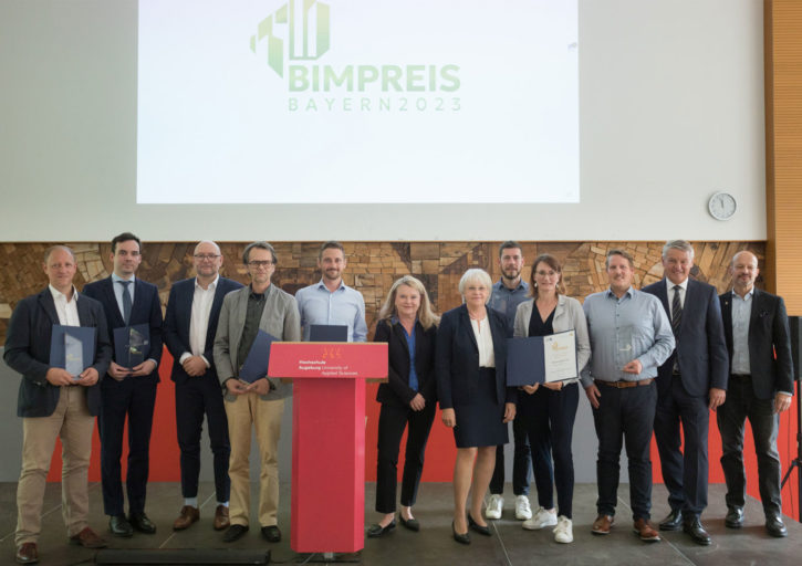 Alle Preisträger:innen und Laudator:innen (12 Personen) der BIM Preise Bayern 2023 stehen auf einer Bühne. Im Hintergrund wird "BIMPREIS Bayern 2023" an die Wand projiziert.