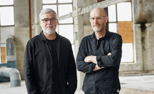 Biwermau Architekten: Zwei Männer mit grauen Haaren/Bärten und schwarzen Klamotten in einer verlassenen Industriehalle