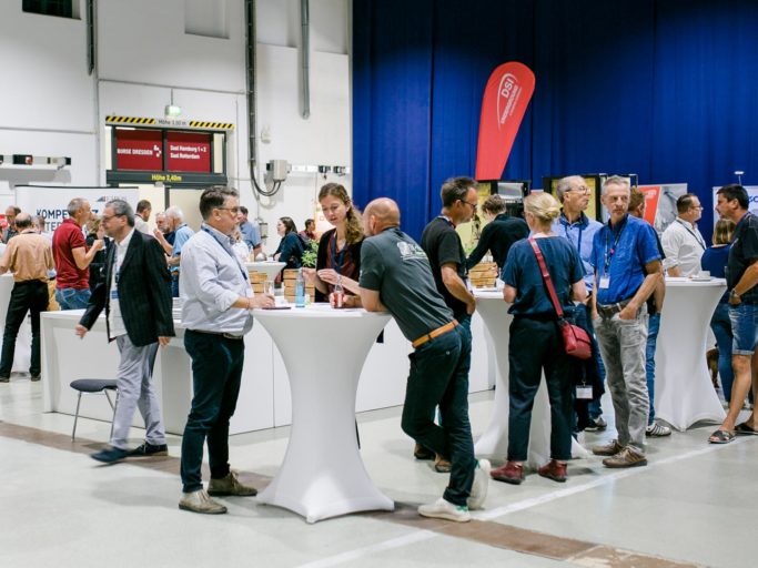 Einige Menschen stehen in einer großen Halle an runden Stehtischen und unterhalten sich.