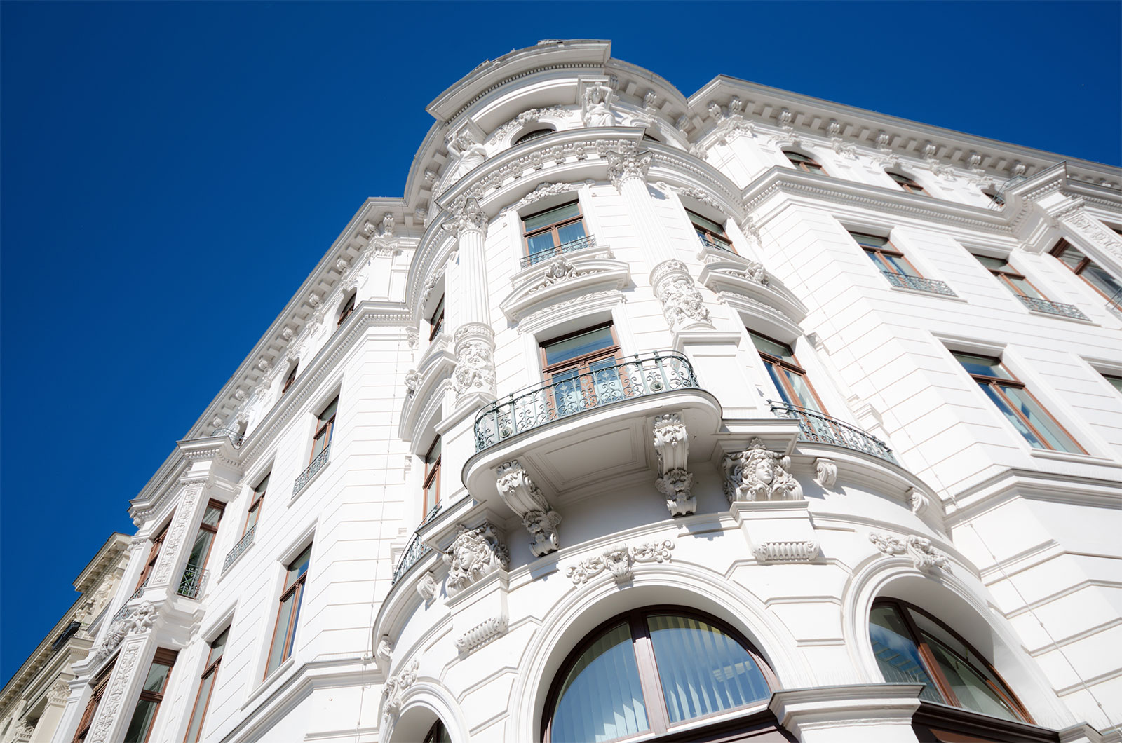 Abgerundete Gebäudeecke eines verzierten Altbaus von unten. Weiße Fassade, braune Fensterrahmen
