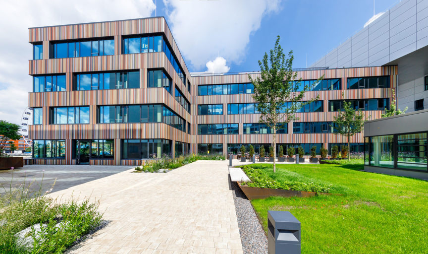 Das Foto des i9-Plaza auf dem iCampus im Werksviertel Münchens: breite Fensterflächen lassen viel tageslicht in das L-förmige Gebäude. Die Fassade wird geprägt von bunten dünnen senkrechten Streifen (hauptsächlich in roten, oragenene und gelben Farbtönen).