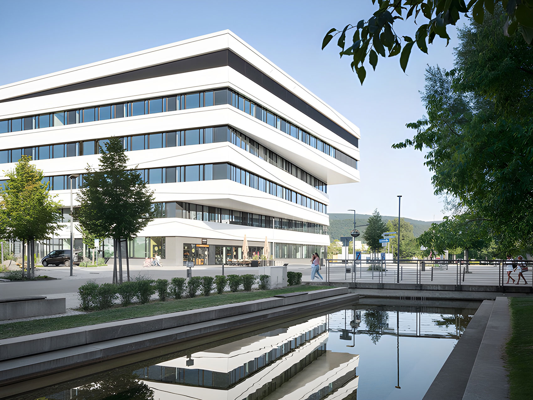 Ein fünfstöckiges Geschäftshaus mit weißer Fassade und langen Fensterstreifen über die ganze Breite spiegelt sich in einem Wasserbecken