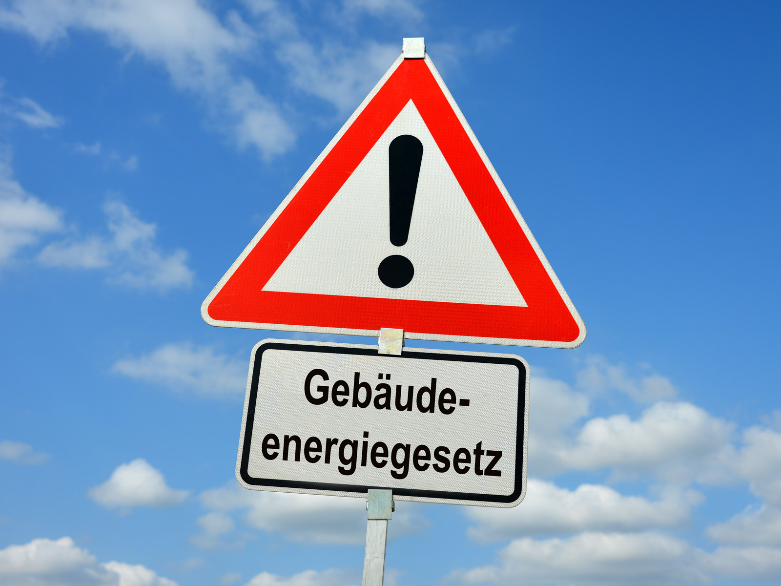 Dreieckiges Straßenschild mit rotem Rand und Ausrufezeichen in der weißen Mitte, darunter ein weiteres Schild mit der Aufschrift: Gebäudeenergiegesetz. Es symbolisiert die angeheizte Debatte um das Gesetz, zu dem viel Kritik geäußert wird.