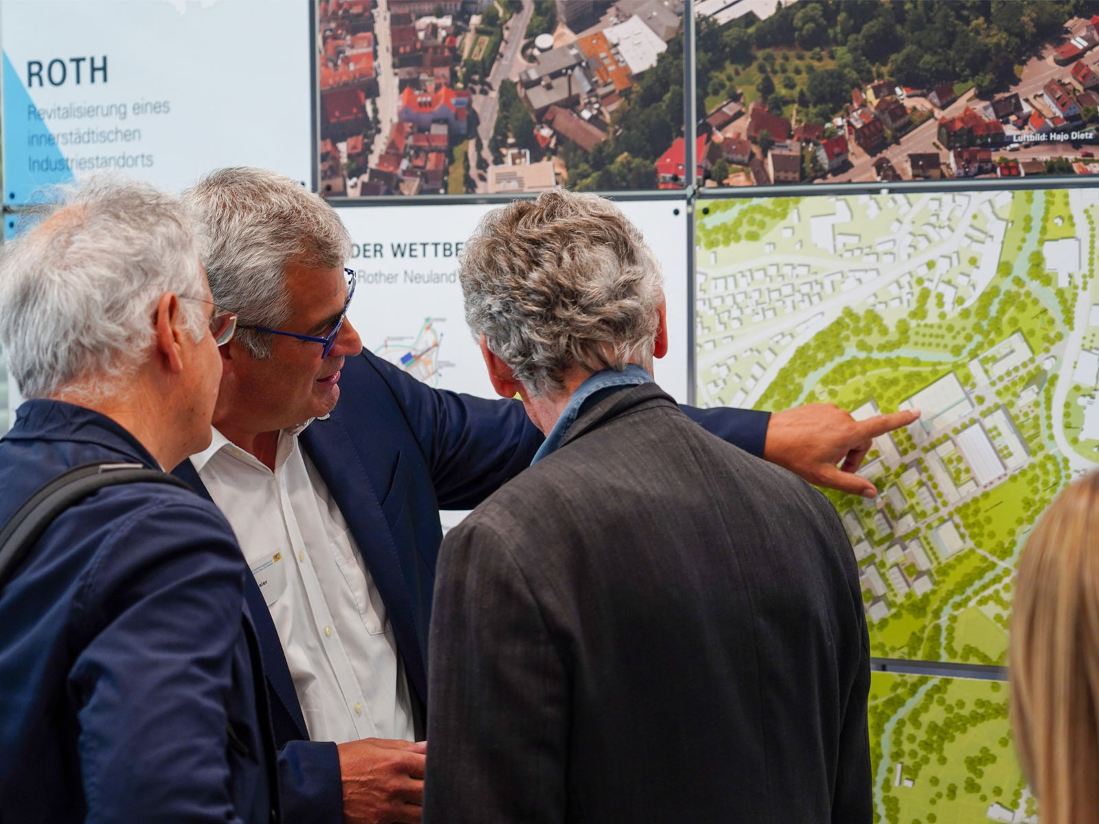 Drei Männer stehen bei der Projektschau für Landstadt Bayern vor einer Werbetafel. Einer von ihnen zeigt auf den abgebildeten Entwurf, während die anderen mit den Blicken seinem Finger folgen.