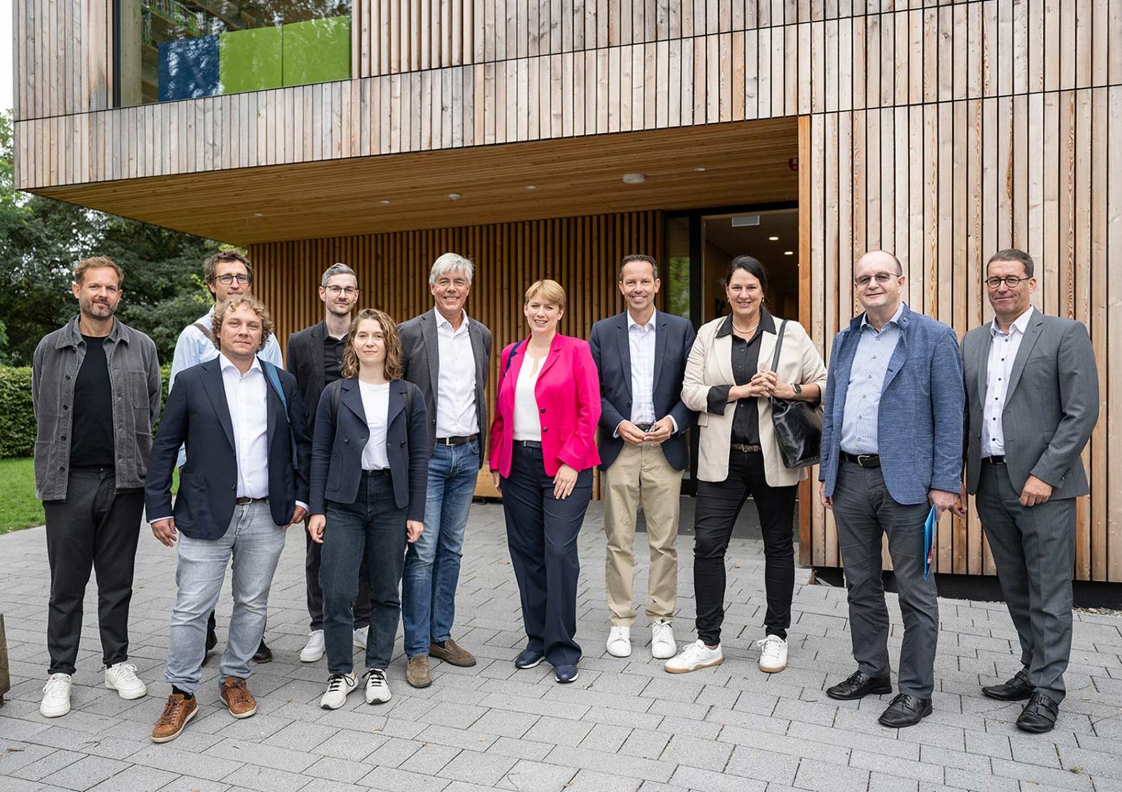 11 Personen (Bürgermeister, Architekten, Ingenieure, ...) stehen vor einem Holzbau-Gebäude. Darunter Staatssekretärin Lindlohr, die im Rahmen von N!BBW (Nachhaltiges Bauen in Ba-Wü) auf Sommertour ist.