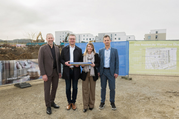 4 Personen stehen vor dem Bauzaun des Projekts Neckar.Rooms im Neckar.Au Viertel. Zwei von ihnen halten eine Zeitkapsel.