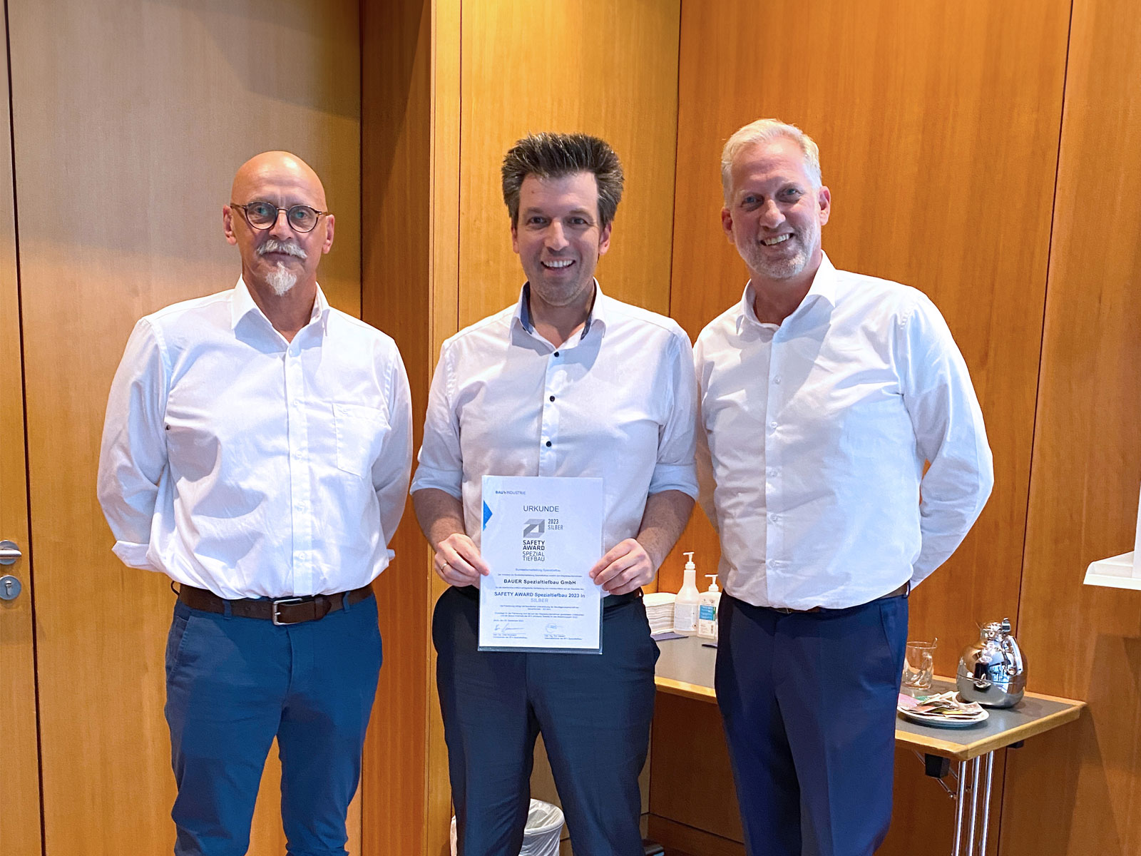Drei Herren der mittleren Alters (Mitarbeiter der BAUER AG) stehen vor einer Holzwand. Der mittlere hält die Urkunde des Safety Awards für Spezialtiefbau in den Händen.