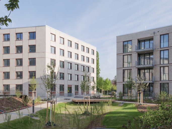 Zwei vier- bis 5-stöckige Gebäude des Neubaus Spinelli in Mannheim. Im Vordergrund ist eine begrünte Fläche mit neu angelegten Pflanz-Flächen zu sehen.
