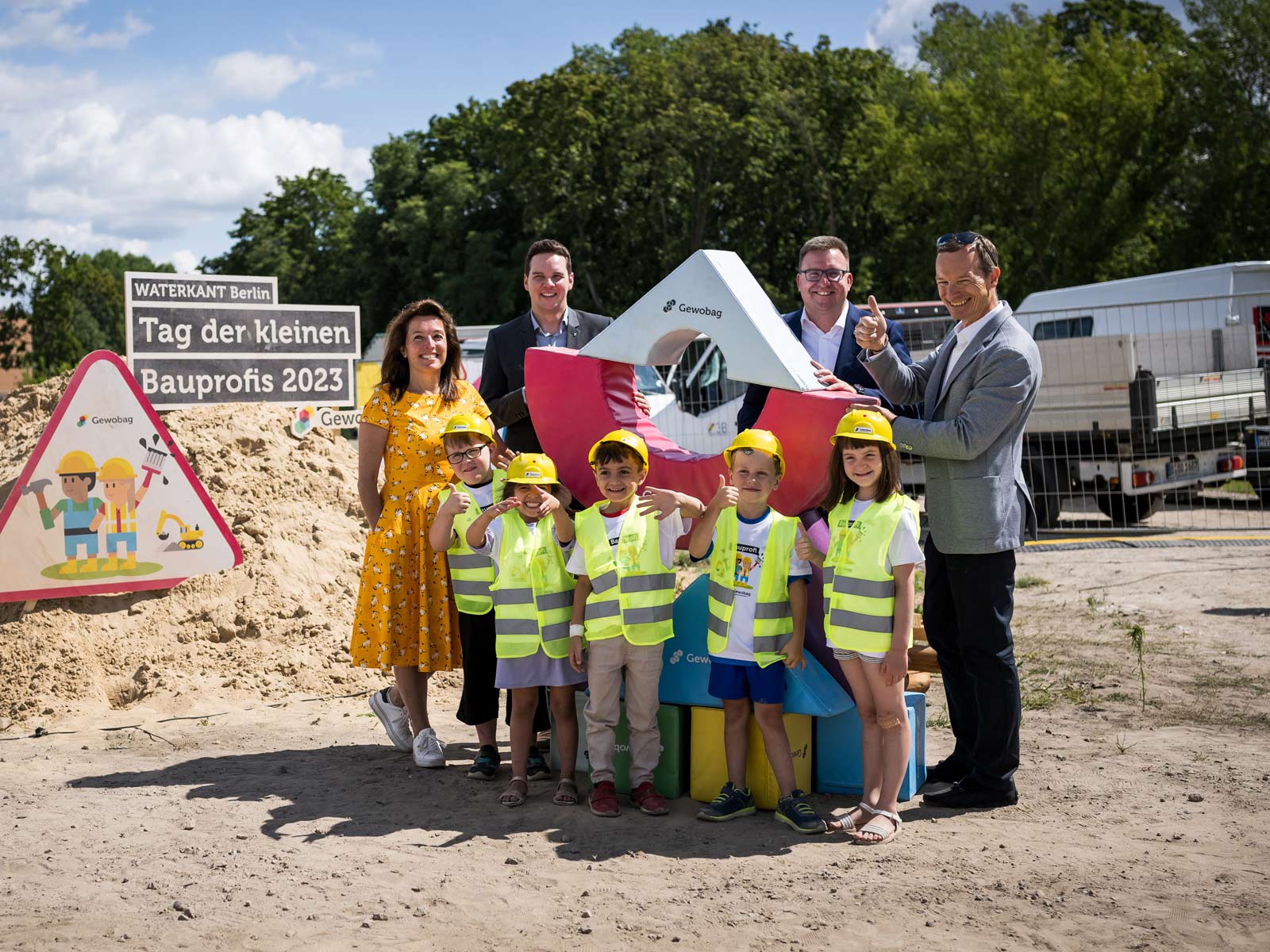 Im Hintergrund ist ein Sandhaufen mit einem Schild "Tag der kleinen Bauprofis 2023". Im Vordergrund sind drei Männer und eine Frau, die ein großes Gewobag-Logo festhalten. Vor ihnen stehen drei grinsende Jungen und zwei Mädchen mit gelben Bauhelmen und neongelben Warnwesten.