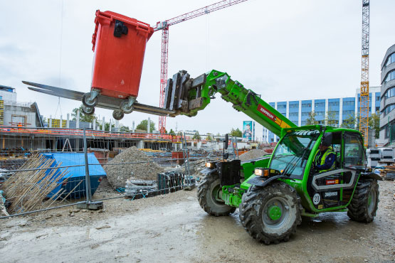 Grünes Baustellenfahrzeug hebt mit Gabeln eine rote Mülltonne hoch