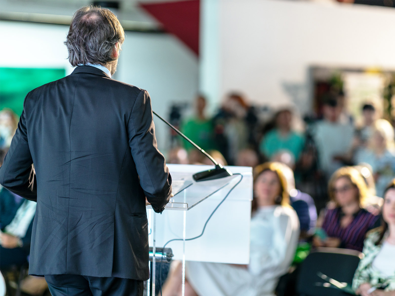 Aus dem Beitragsbild zum Zukunft Bau Kongress 2023 ist ein Mann in Anzug von hinten zu sehen. Er steht an einem Rednerpult. Menschen sitzen vor ihm und hören ihm zu. Die Kamera blickt in Richtung der Zuschauer.